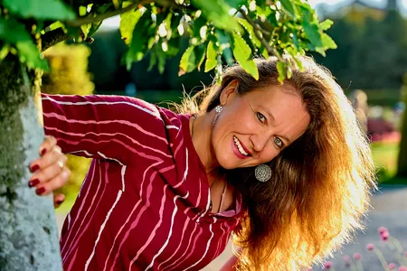 Eine Frau mit langen Haaren und einem rot gestreiften Hemd lehnt lächelnd an einem Baum. Der sonnenbeschienene Hintergrund besteht aus Grünpflanzen und einem unscharfen Weg.